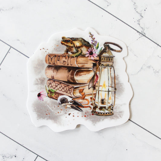 vinyl sticker featuring watercolor design of stacked spell books with frog on top, lantern, and other magical elements