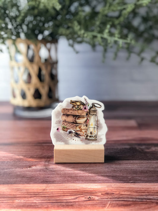 vinyl sticker featuring watercolor design of stacked spell books with frog on top, lantern, and other magical elements