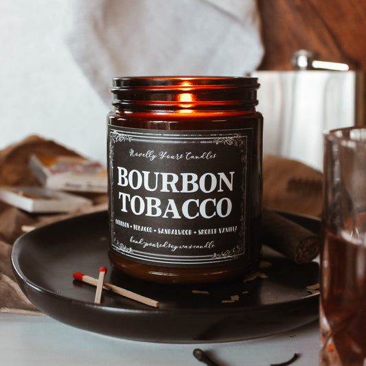 bourbon tobacco named candle in amber glass jar with black lid, sits on top of black tray surrounded by wood, matches, and cigar