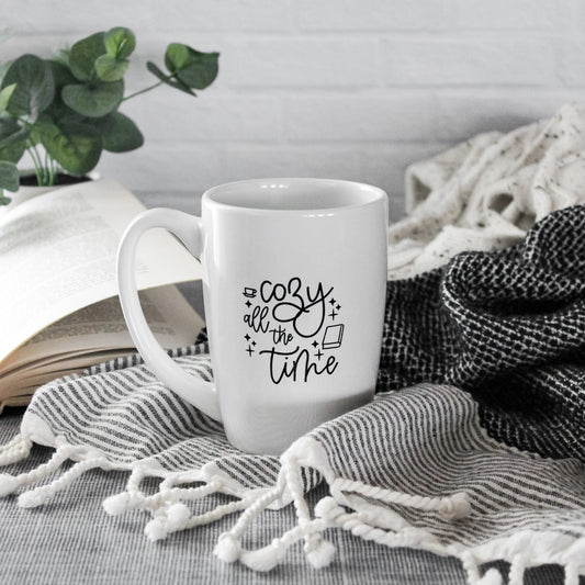 white mug with black script text reading "Cozy all the time" with graphics of a coffee mug & book