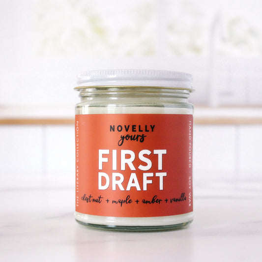 bookish candle "First Draft" with rusty orange label sits in front of clean kitchen background on marble countertop