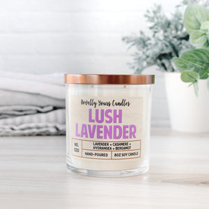 lush lavender scented soy wax candle in clear glass tumbler with bronze lid. Sits on beechwood surface with white brick background framed by other home goods