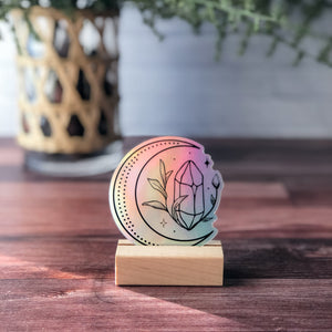 shiny holographic moon & crystal sticker with rainbow iridescent finish. sticker design is crescent moon black outline with crystal, stars, and leafy accents