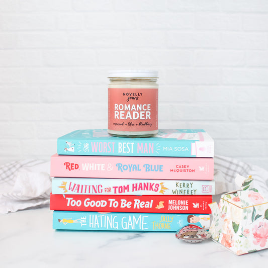 romance reader scented soy wax candle in clear glass jar with white lid and soft rose colored label on top of pink and blue book stack of romance books
