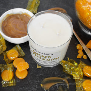 smoked butterscotch scented candle with butterscotch colored text. candle is in clear glass tumbler jar and sits on black surface from the top view down. Two wicks are visible. Candle is surrounded by butterscotch candies and sauce