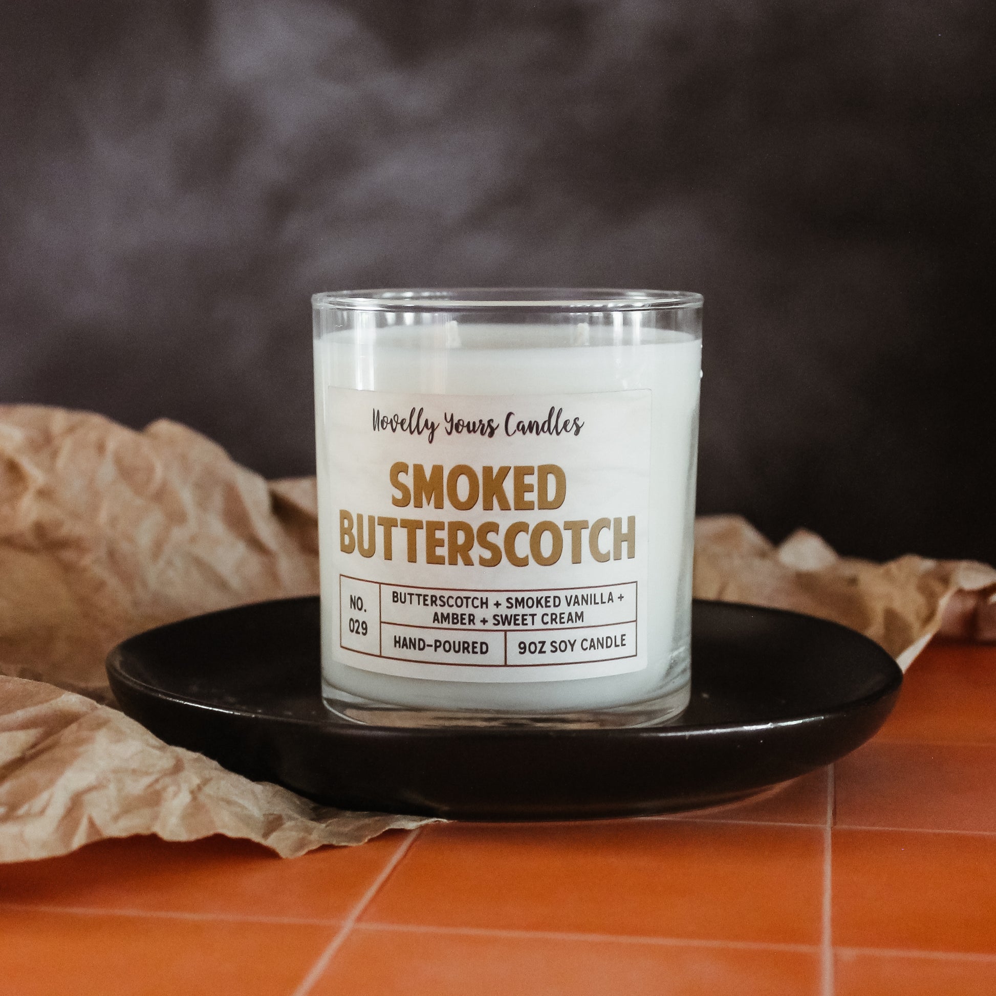 smoked butterscotch scented candle with butterscotch colored text. candle is in clear glass tumbler jar with lid off. Candle sits on round black plate which is on top of orange tiled countertop with smoky black background. brown Kraft paper accents the scene.