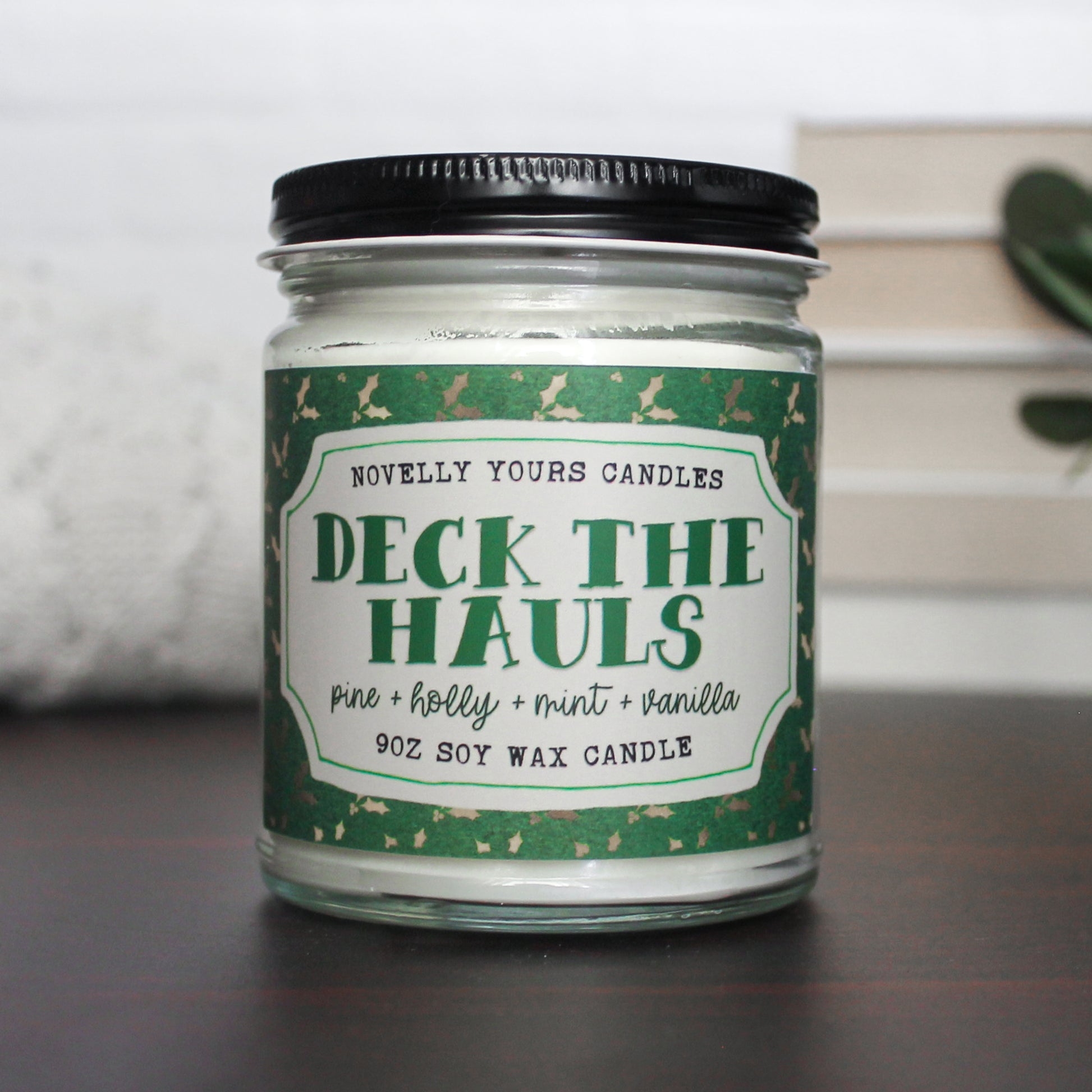 deck the hauls book themed christmas candle inspired by book presents and gifts. candle is white wax in clear jar with green label showing holly in gold and has a black metal lid