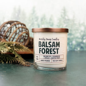 scented candle with balsam forest title in clear jar with bronze lid against pine tree background