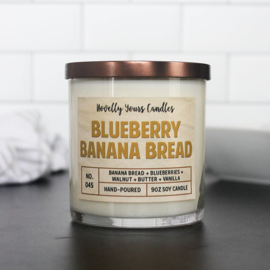 blueberry banana bread candle with text in a dark yellow text. candle has bronze flat lid and is in clear glass jar, sits on top of black surface with white tile background