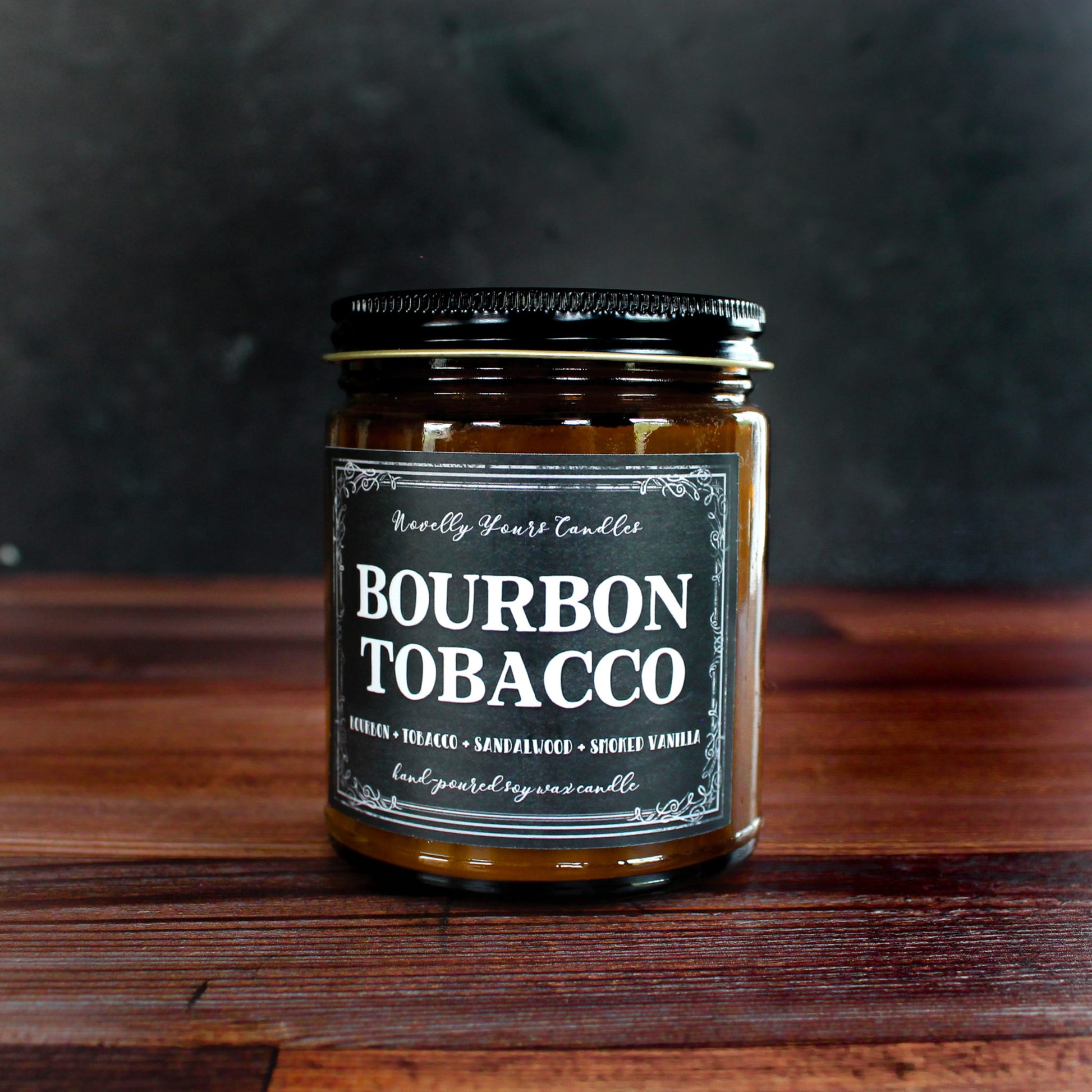bourbon tobacco named candle in amber glass jar with black lid, sits on top of wood background