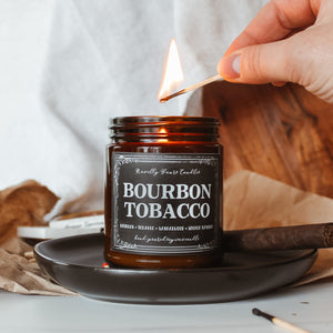bourbon tobacco named candle in amber glass jar with black lid, sits on top of black tray surrounded by wood, matches, and cigar
