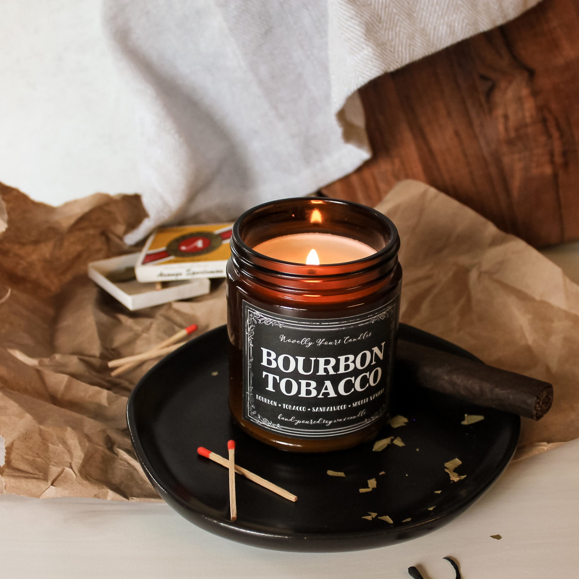 bourbon tobacco named candle in amber glass jar with black lid, sits lit on black tray surrounded by cigar, matches, and tobacco leaves