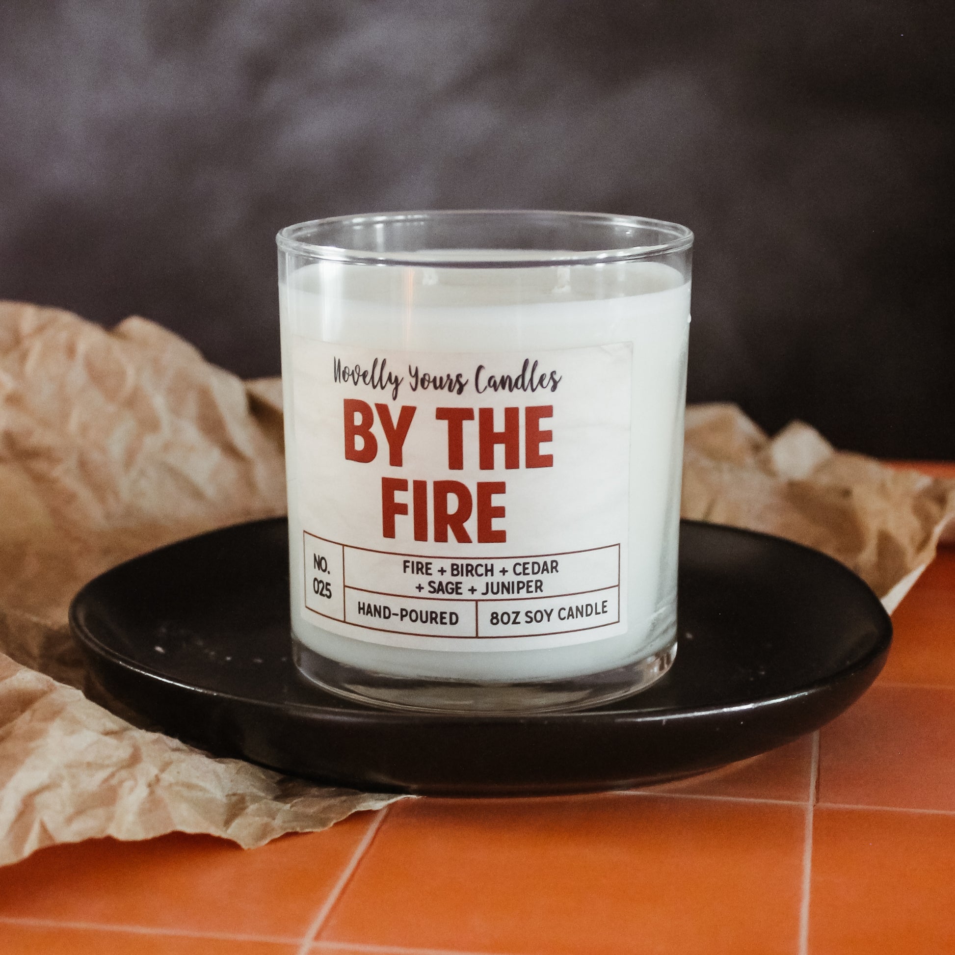 by the fire scented soy wax candle in glass tumbler. sits on black plate on top of peach tile countertop
