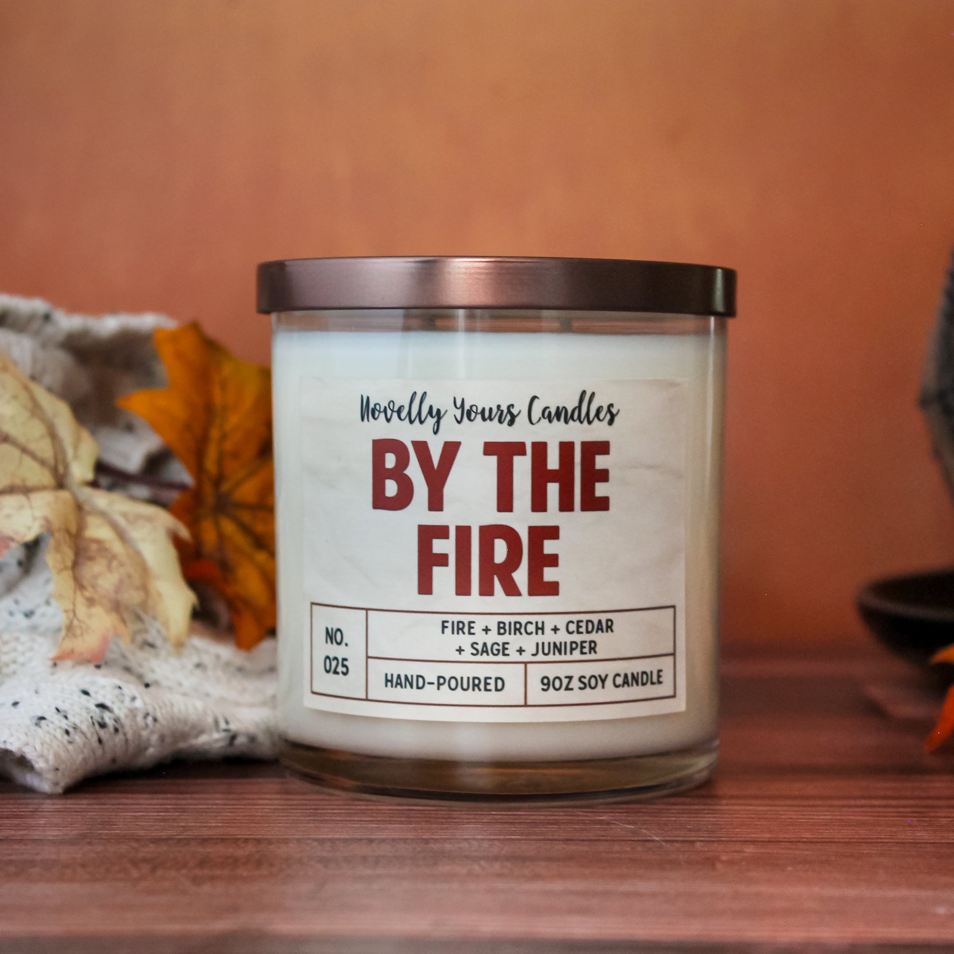 by the fire scented soy wax candle in glass tumbler and bronze lid. Sits on wooden surface with orange background