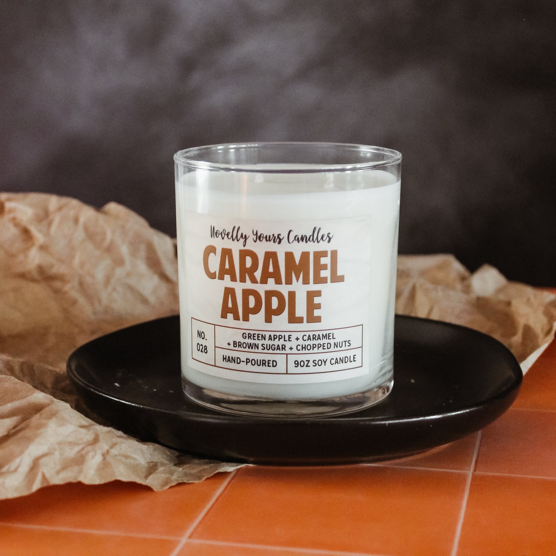 caramel apple scented candle by novelly yours, sits on round black plate on top of peach tiled counter. Set against smoky black background and accented with brown paper. Candle is in clear glass tumbler jar with lid off