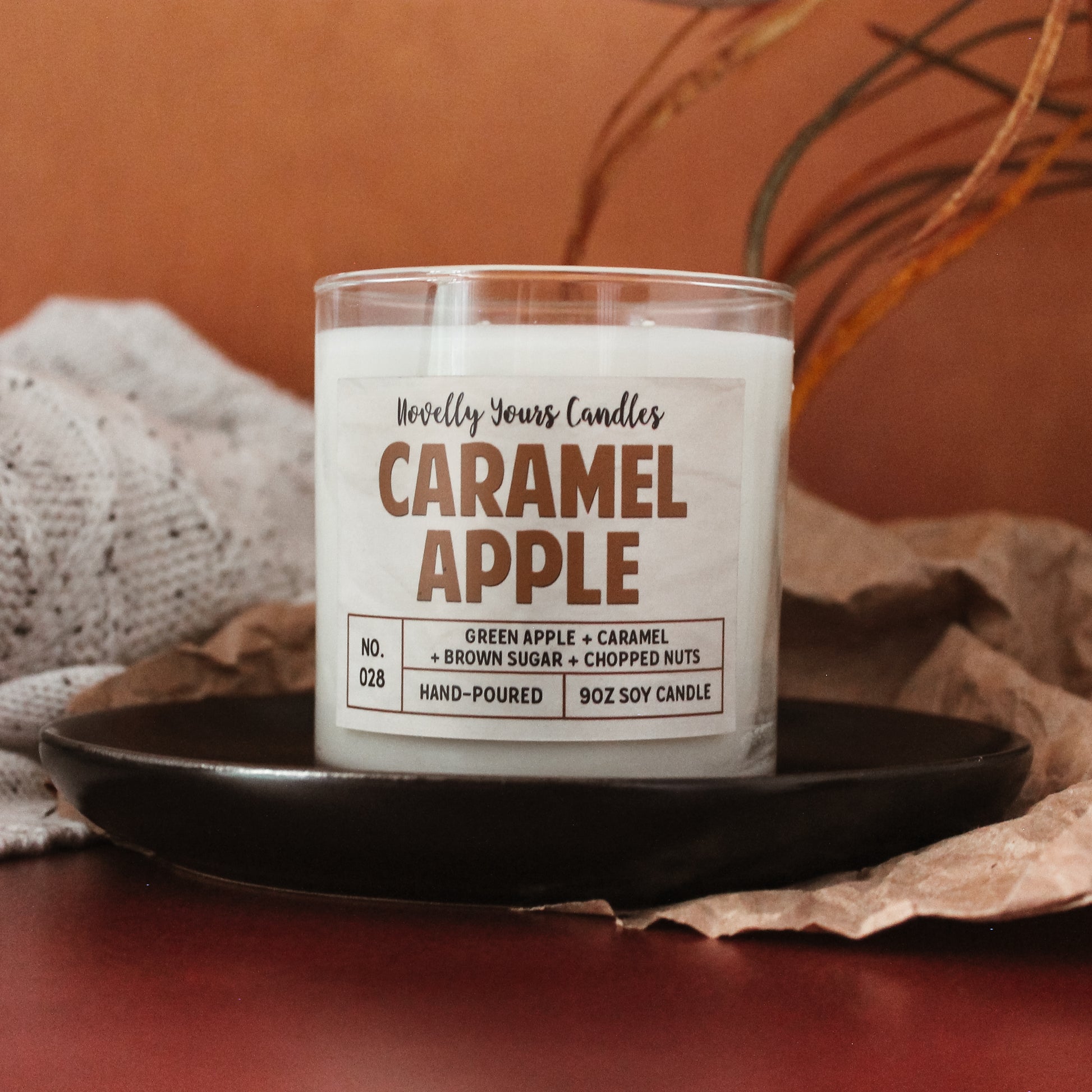 caramel apple scented candle by novelly yours, sits on round black plate on top of rustic red surface. Set against burnt orange background and accented with brown paper. Candle is in clear glass tumbler jar with lid off