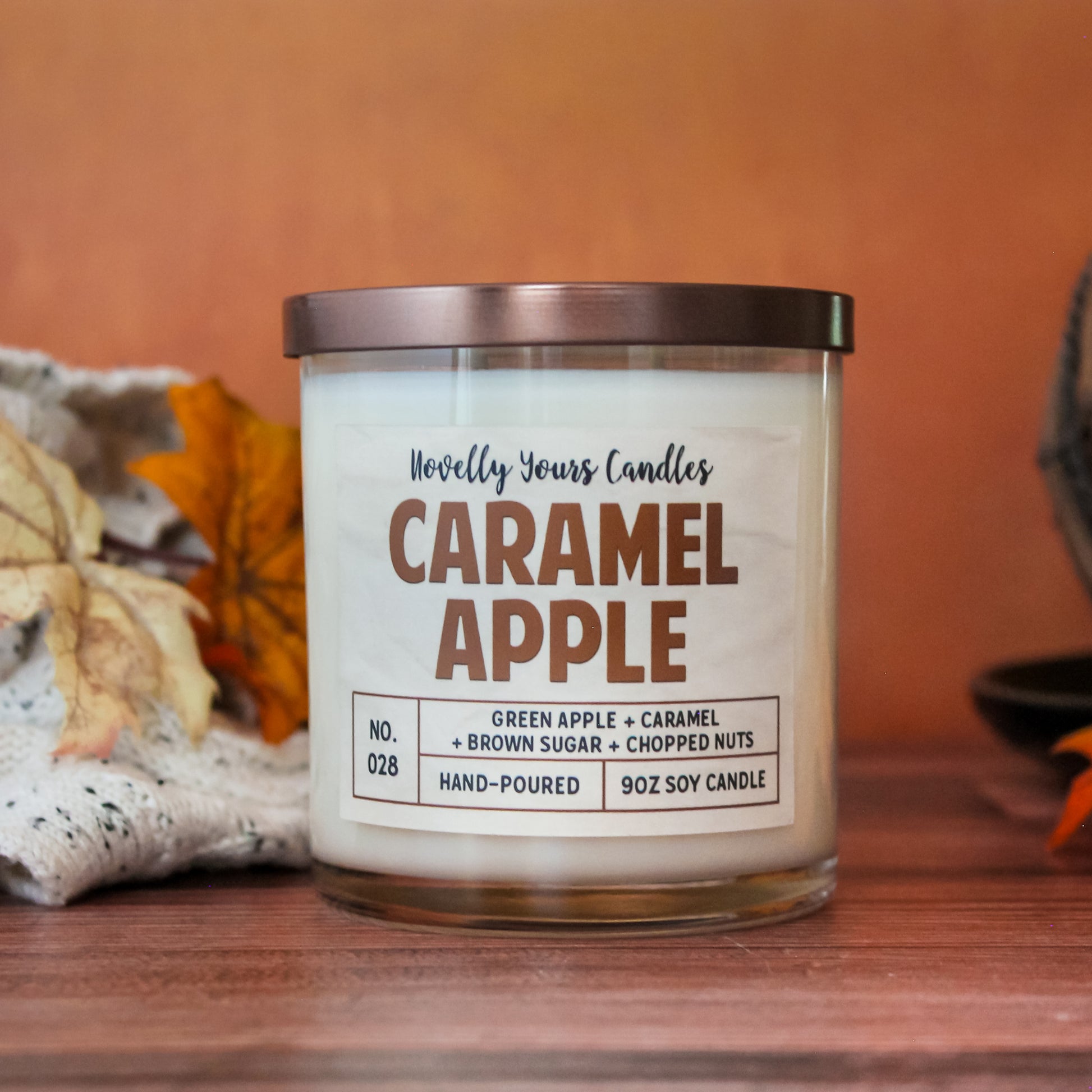 caramel apple scented candle by novelly yours, sits on round black plate on top of wooden surface. Set against orange background. Candle is in clear glass tumbler jar with lid off
