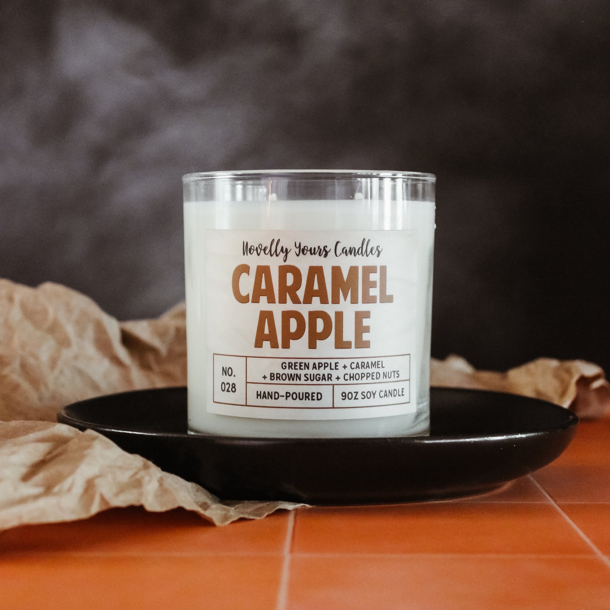 caramel apple scented candle by novelly yours, sits on round black plate on top of peach tiled counter. Set against smoky black background and accented with brown paper. Candle is in clear glass tumbler jar with lid off