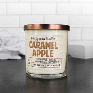caramel apple scented candle by novelly yours. candle is in clear glass tumbler jar with bronze lid and caramel colored text.