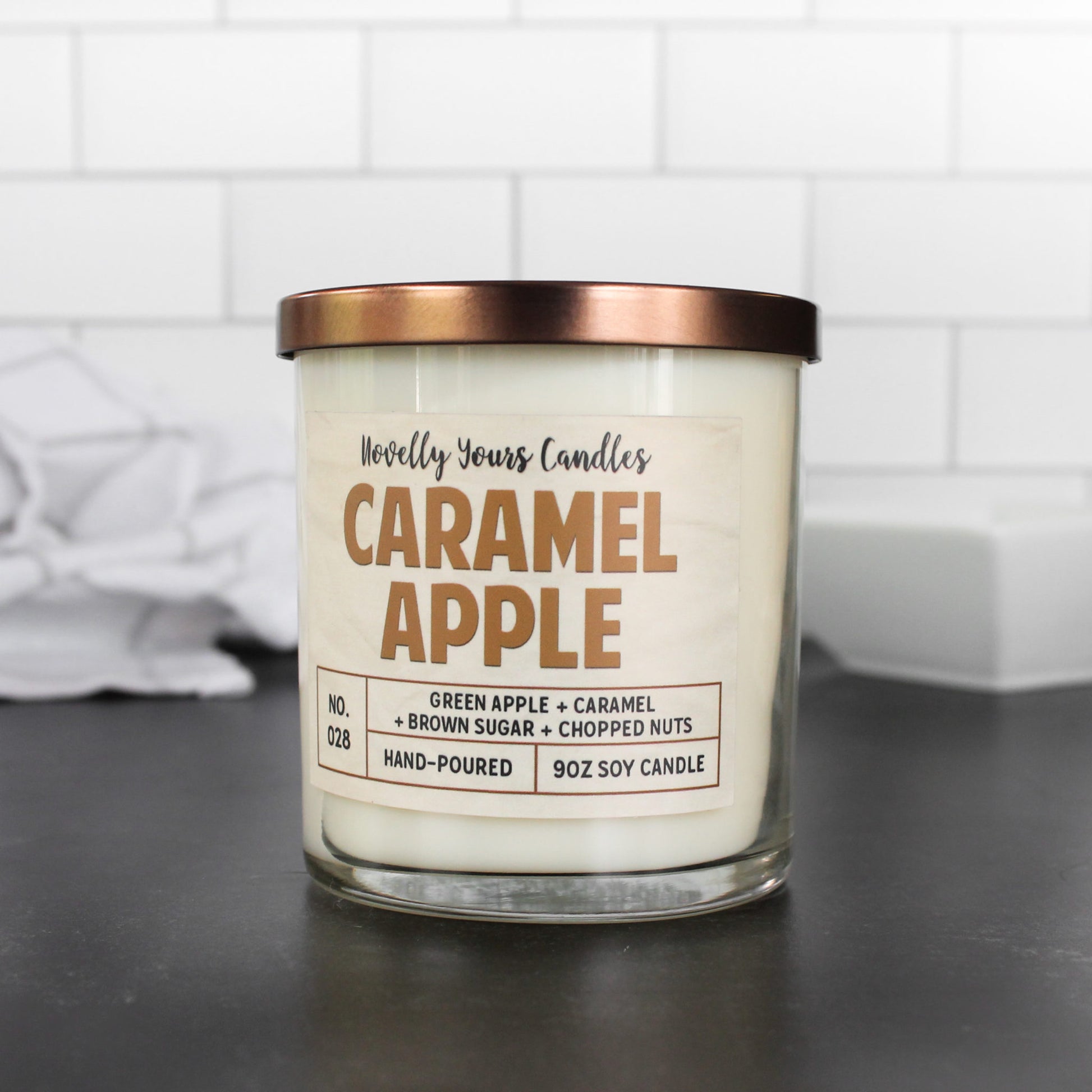 caramel apple scented candle by novelly yours. candle is in clear glass tumbler jar with bronze lid and caramel colored text.