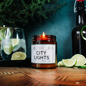 city lights scented soy wax candle in amber jar, lit surrounded by limes & cocktails