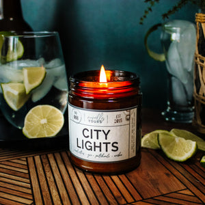 city lights scented soy wax candle in amber jar, lit surrounded by limes & cocktails