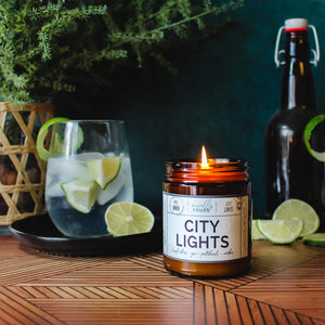 city lights scented soy wax candle in amber jar, lit surrounded by limes & cocktails