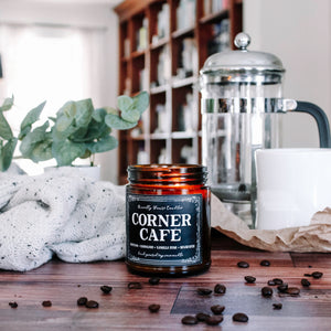 corner cafe scented soy wax candle in amber jar, lit on wooden background surrounded by coffee beans and cozy items