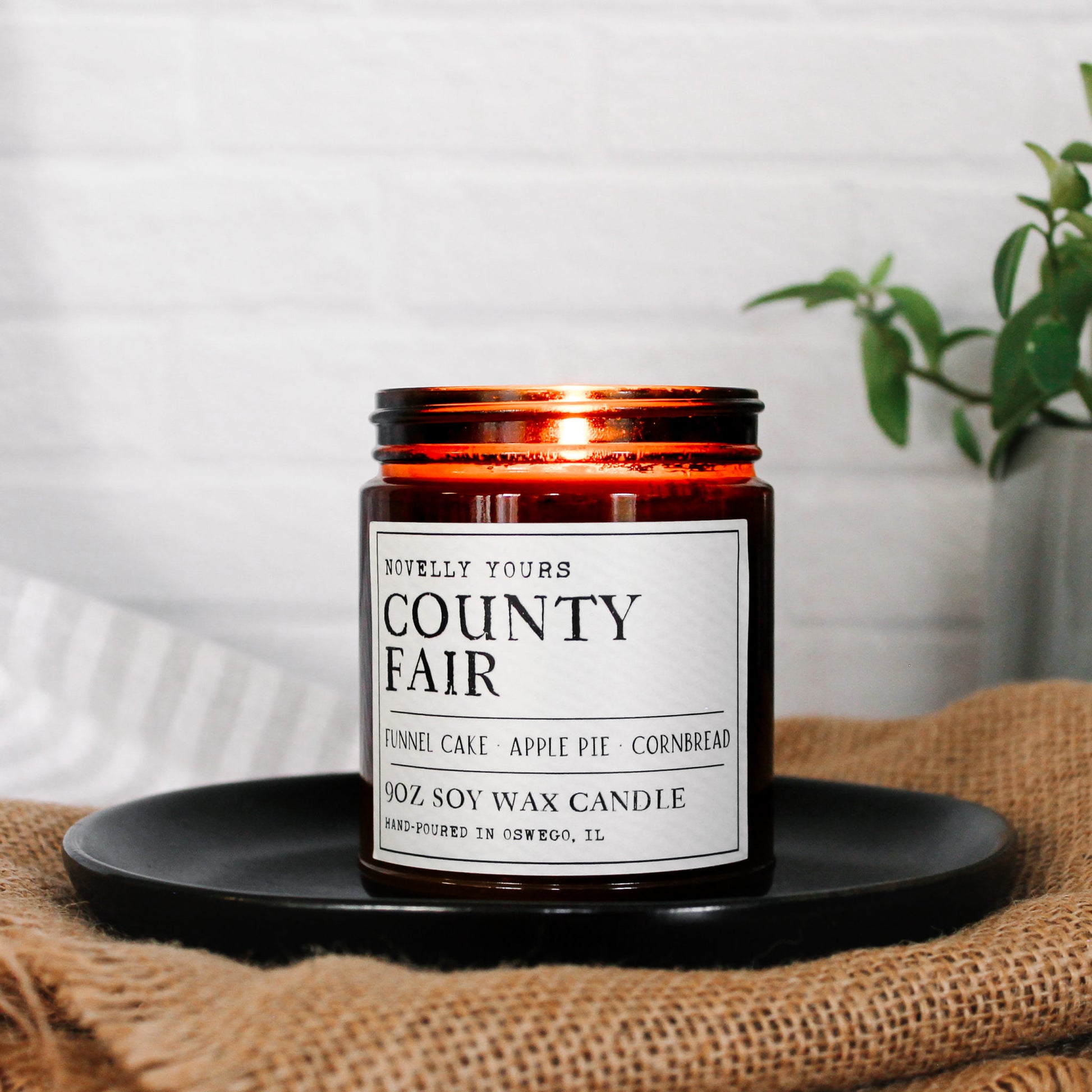 county fair bakery scented soy wax candle in amber jar, sits on black plate and burlap with white background