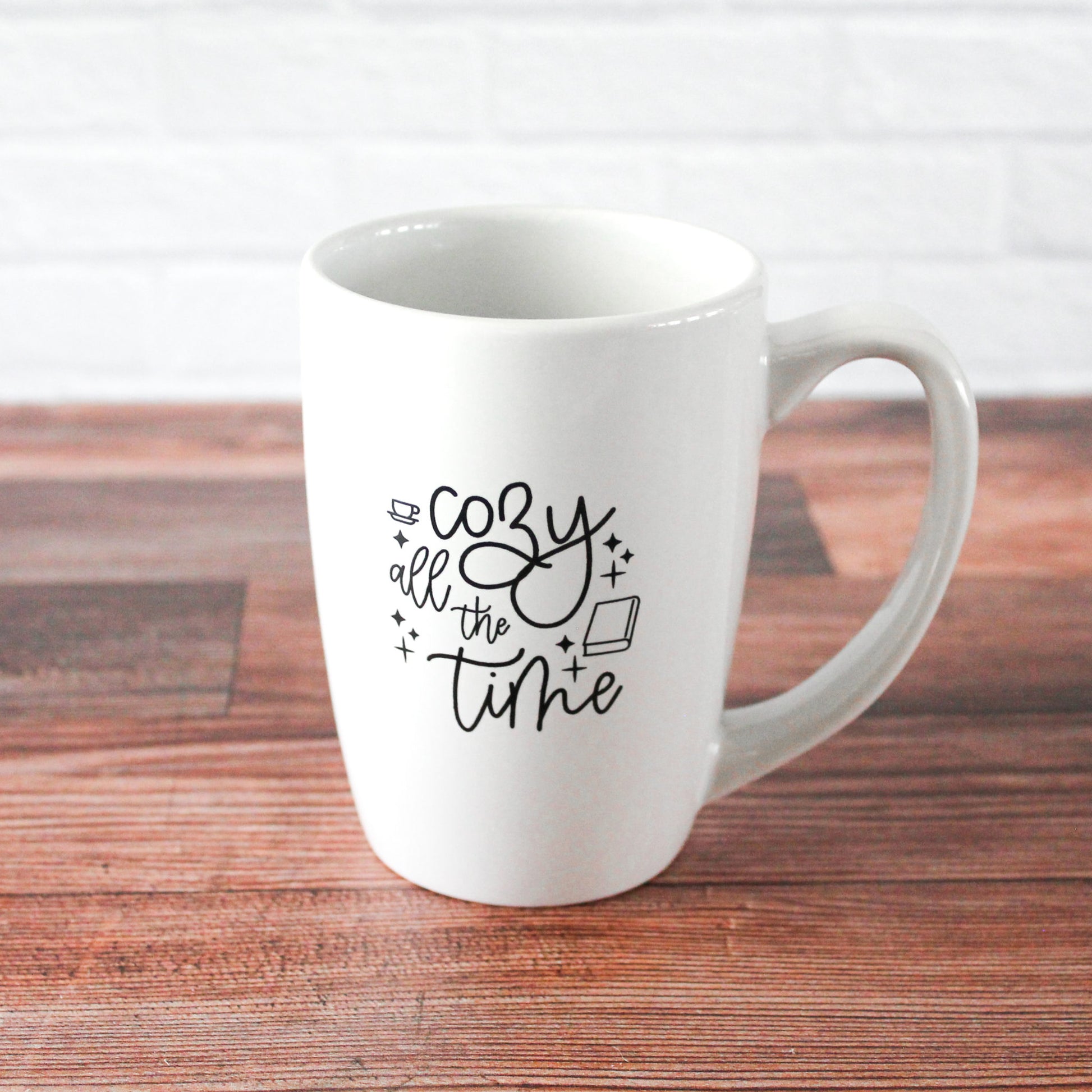 white mug with black script text reading "Cozy all the time" with graphics of a coffee mug & book