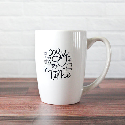 white mug with black script text reading "Cozy all the time" with graphics of a coffee mug & book