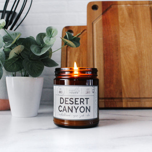 desert canyon scented soy wax candle in amber jar, lit on marble surface in front of kitchen items