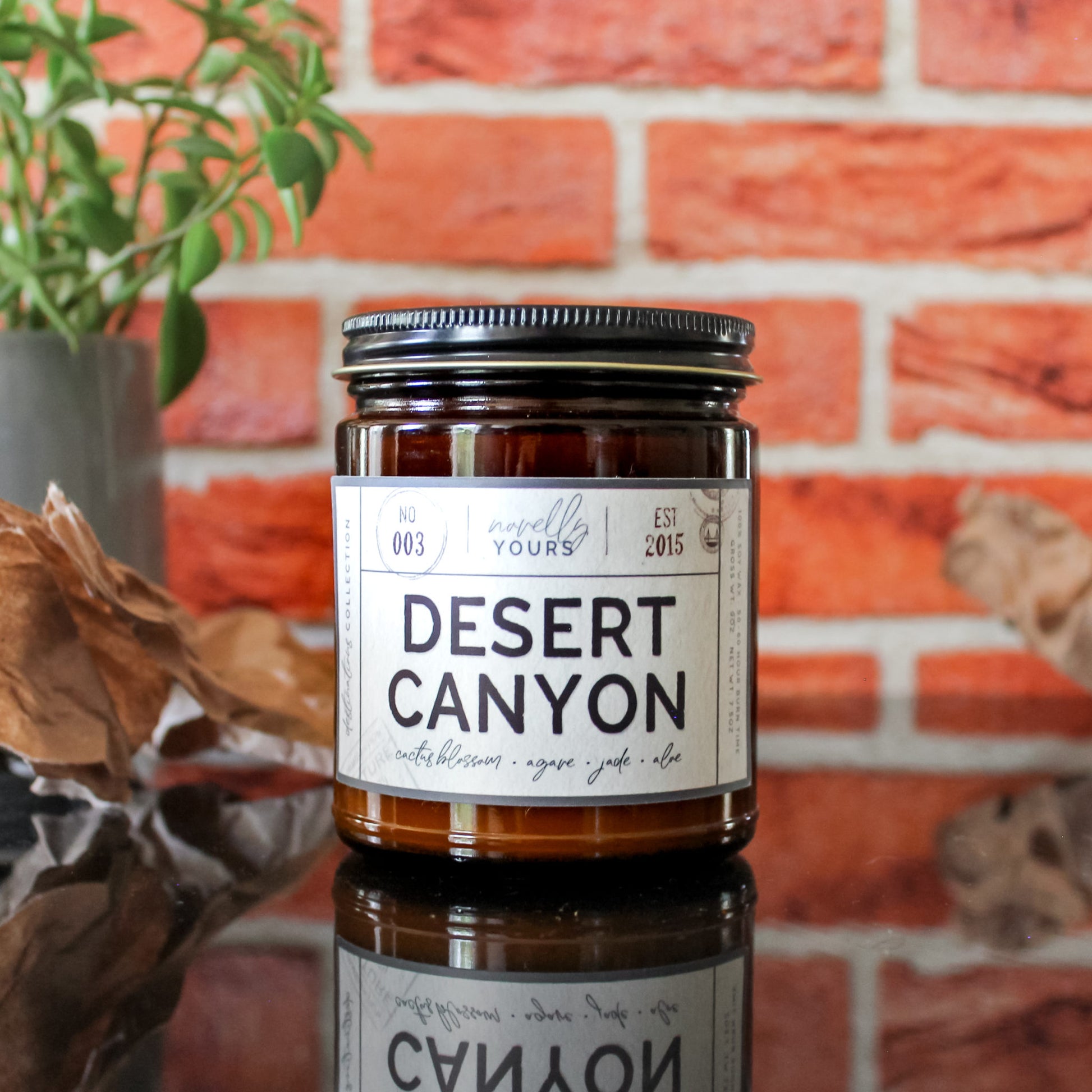 desert canyon scented candle in amber jar with black lid, sits on black reflective base with red brick background