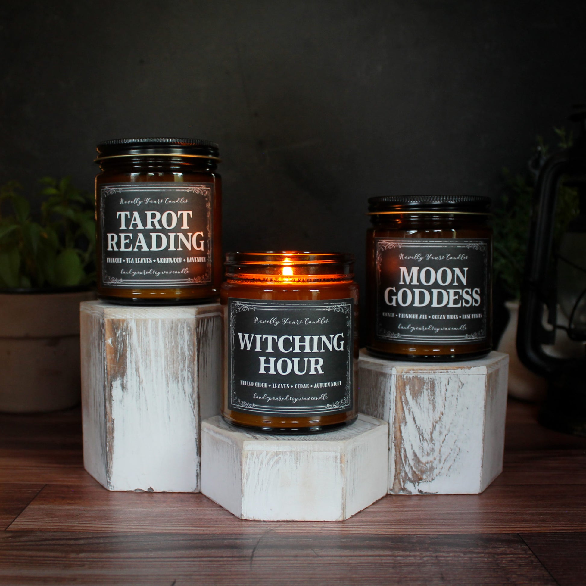three Novelly Yours candles: tarot reading, witching hour, and moon goddess. Each sits on a hexagonal white wooden pedestal, all of different heights. Pedestals rest on wooden surface with black background and dark, moody aesthetic.