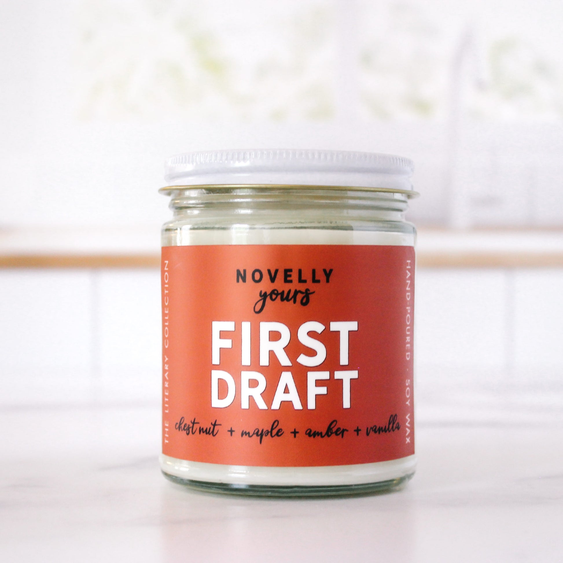bookish candle "First Draft" with rusty orange label sits in front of clean kitchen background on marble countertop