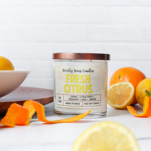 fresh citrus soy wax candle with yellow text in clear glass tumbler with bronze lid. surrounded by oranges and lemons.