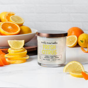 fresh citrus soy wax candle with yellow text in clear glass tumbler with bronze lid. surrounded by oranges and lemons.