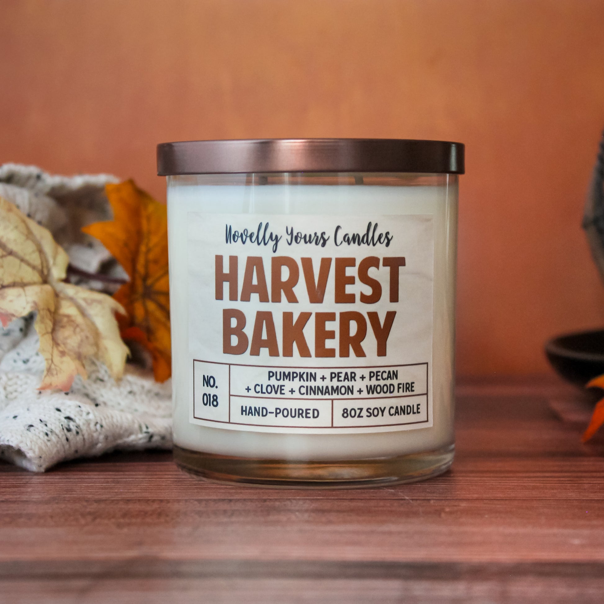 harvest bakery soy wax scented candle in glass tumbler jar with bronze lid. Candle sits on wooden surface against orange background