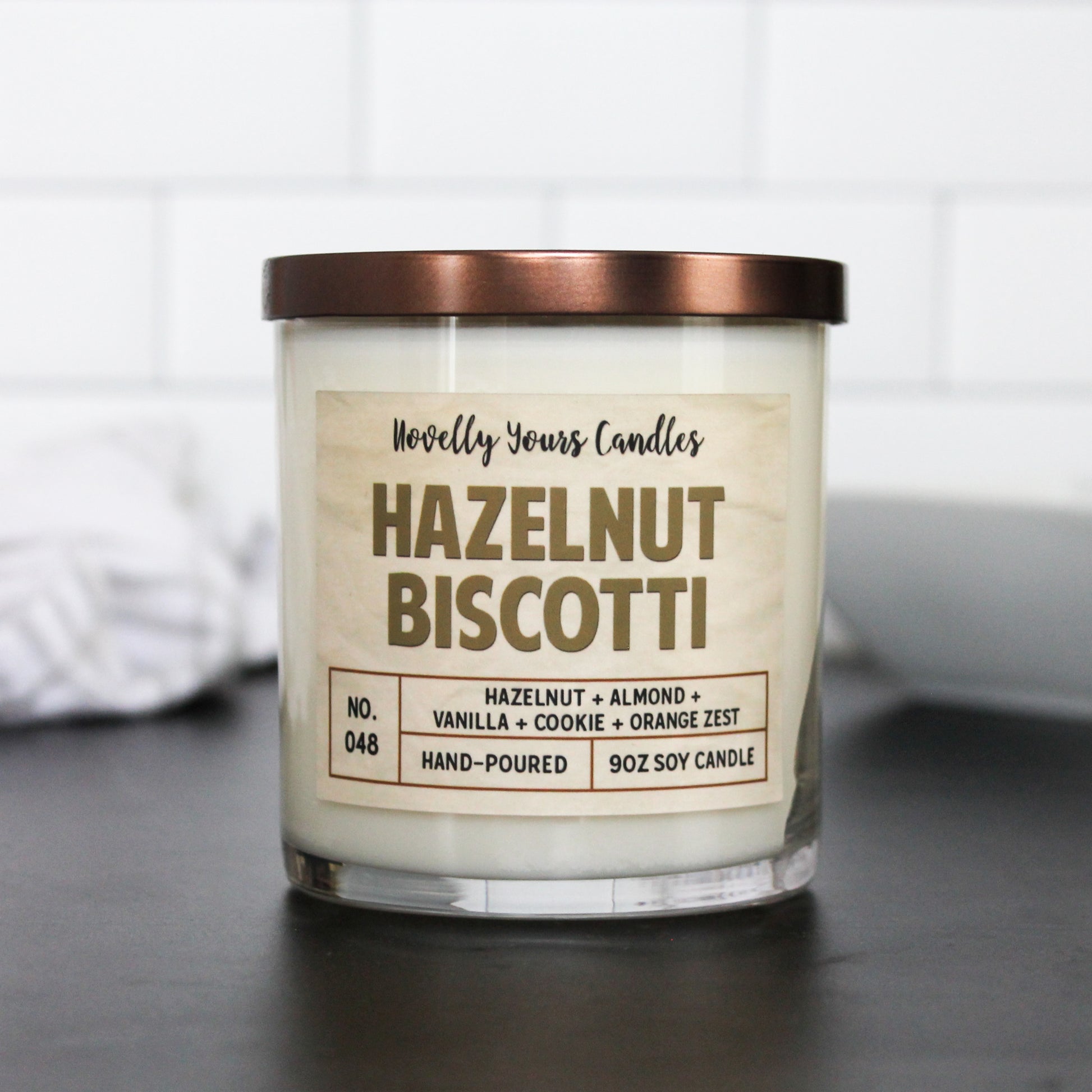 hazelnut biscotti named candle with title in dark tan. candle has bronze flat lid and is in clear jar, which sits on black surface with white tile background