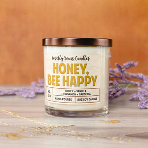 honey bee happy candle in gold honey colored text, candle in glass tumbler jar with bronze lid. sits on wooden surface with honey drizzle and purple flowers