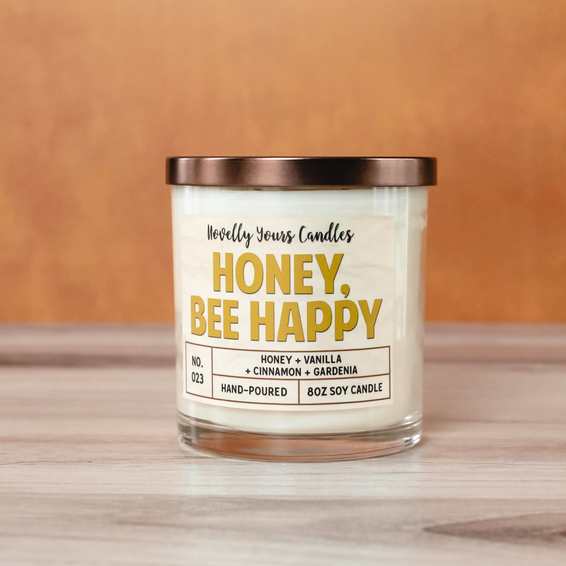 honey bee happy candle in gold honey colored text, candle in glass tumbler jar with bronze lid. Sits on light wood surface with orange background