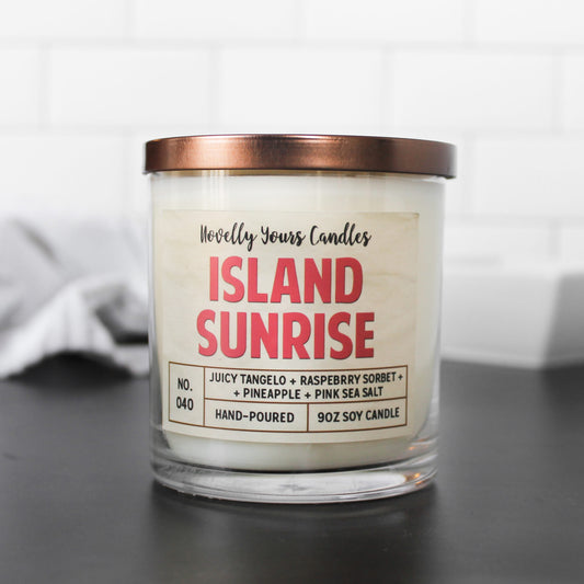 white soy wax candle in straight sided clear tumbler jar with bronze lid featuring candle title "island sunrise" in hot pink block letters. candle sits on black surface with white background