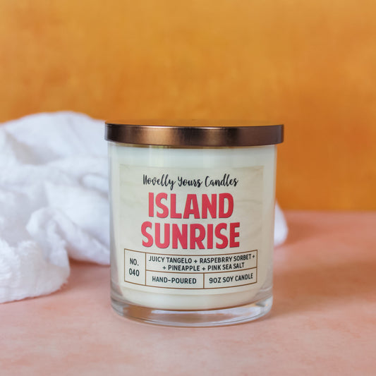 white soy wax candle in straight sided clear tumbler jar with bronze lid featuring candle title "island sunrise" in hot pink block letters. candle sits on pink surface with orange background