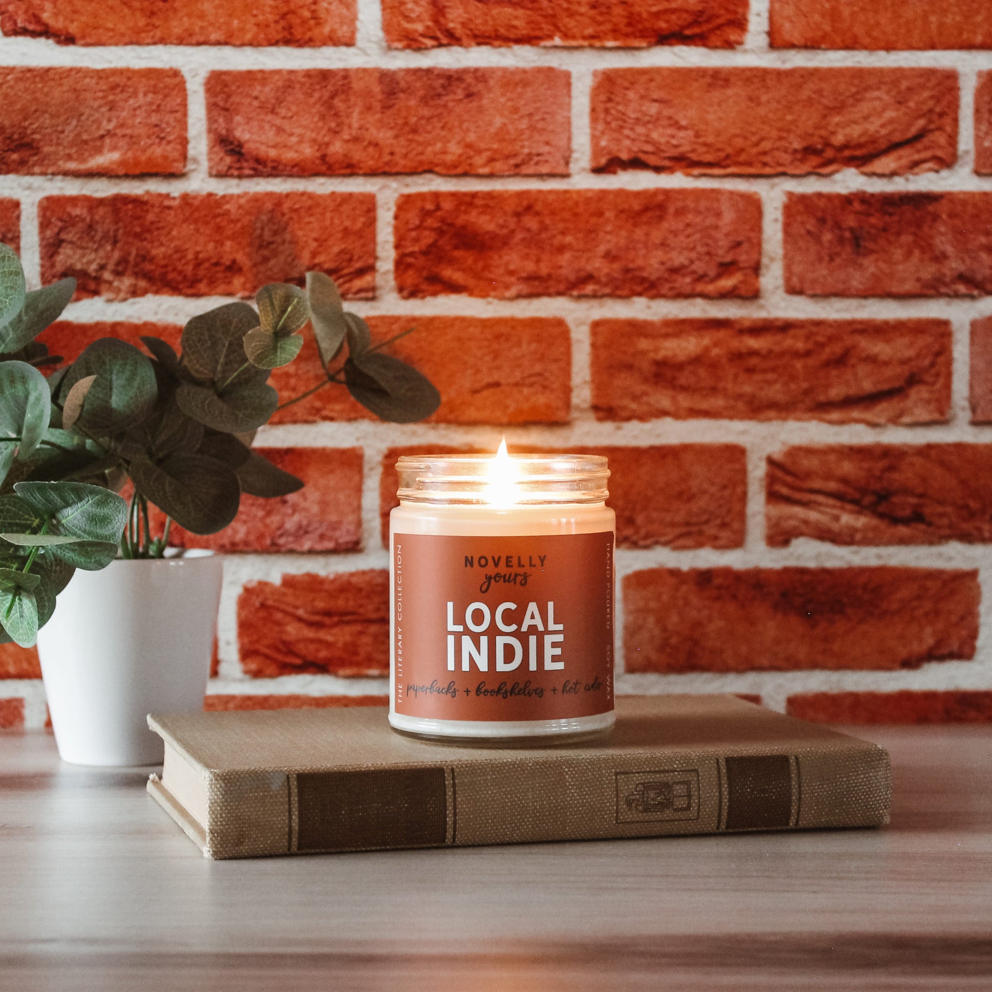 Local indie scented soy wax candle with burnt orange label in clear glass jar with white lid. sits on top of old book, candle is lit, all against red brick background