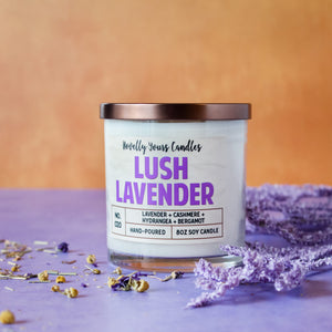 lush lavender scented soy wax candle in clear glass tumbler with bronze lid. Sits on purple surface with orange background.