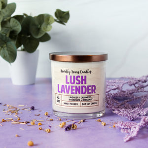 lush lavender scented soy wax candle in clear glass tumbler with bronze lid. Sits on purple surface with soft white background, surrounded by purple flowers and lavender potpourri