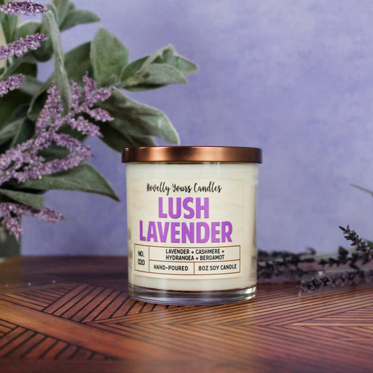lush lavender scented soy wax candle in clear glass tumbler with bronze lid. Sits on mid century style wooden surface with purple background.