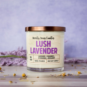 lush lavender scented soy wax candle in clear glass tumbler with bronze lid. Sits on light beechwood surface with purple background