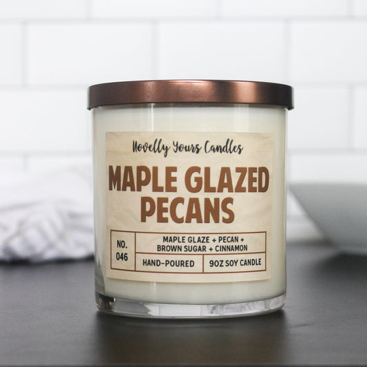 maple glazed pecans candle with caramel colored title text. candle in clear glass jar with flat bronze lid sits on black surface with white tile background