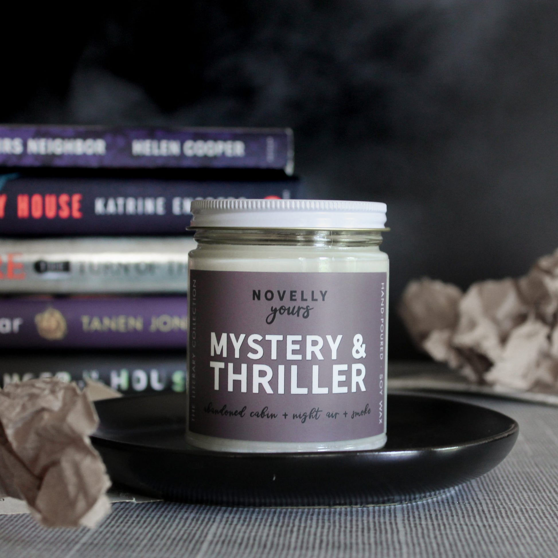 mystery & thriller scented soy wax candle with purple grey label and white lid sits on black round tray with paper accents and stack of thriller books in background